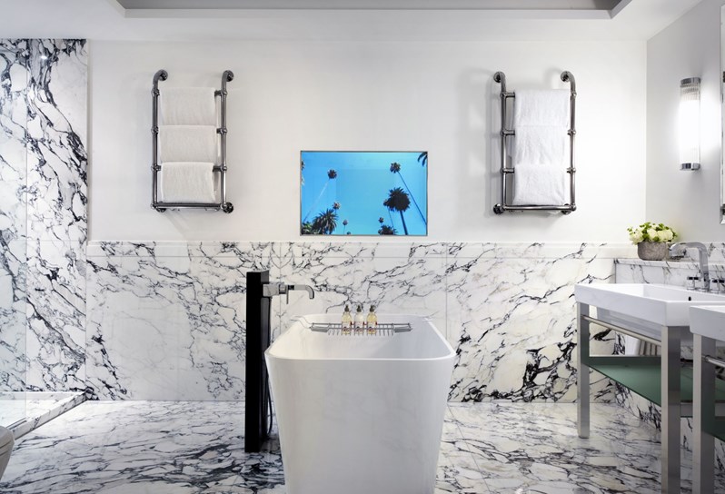A deep soaking tub in the middle of a bathroom with white Carrera marble. On the far wall is a television and two heated towel racks on either side. To the left is the entrance to the standing shower and to the right of the bathtub are two basins and flowers.