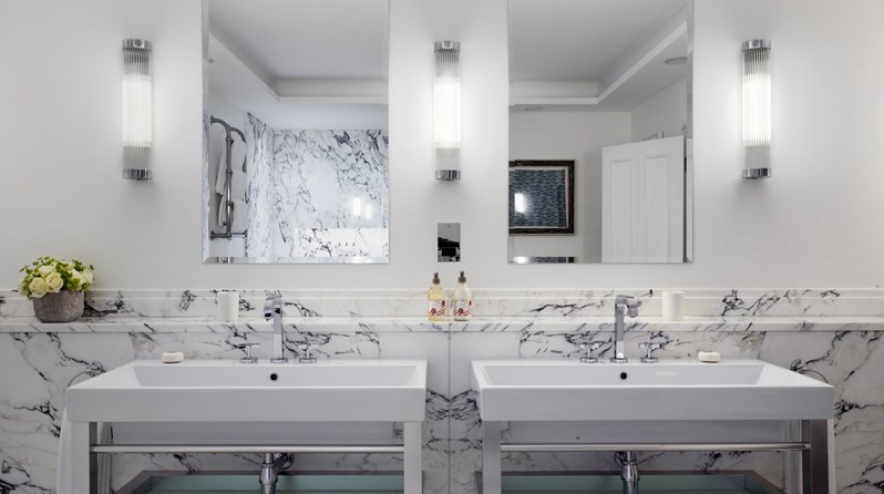 Looking on to two basins in a white Carrera marbled bathroom. There are two mirrors, one above each. Rik Rak products sit on the counter and a flower arrangement is on the left side.