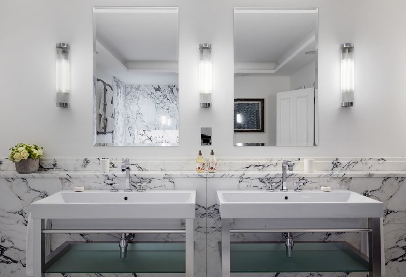 Looking on to two basins in a white Carrera marbled bathroom. There are two mirrors, one above each. Rik Rak products sit on the counter and a flower arrangement is on the left side.