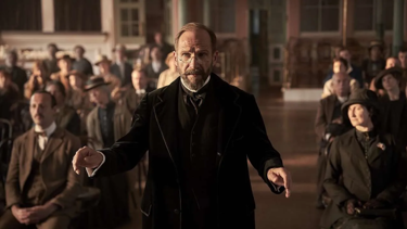 A still from 'The Choral'. An older bearded man dressed in black formal attire from early 1900s and small spectacles, stands in the middle of the shot. Behind him, rows of people can be seen sat down in a large hall.