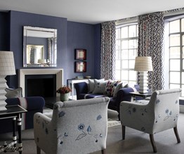 A wide view of the living space in a suite. There are many shades of blue. The walls are a blue grey and the couches face each other and are blue and white vertical stripes. The curtains are in a rik rak pattern. There is a fireplace with a mirror on the wall above and two small pieces of art to the right. The two chairs in the foreground are grey with beautiful flower stitching detail.