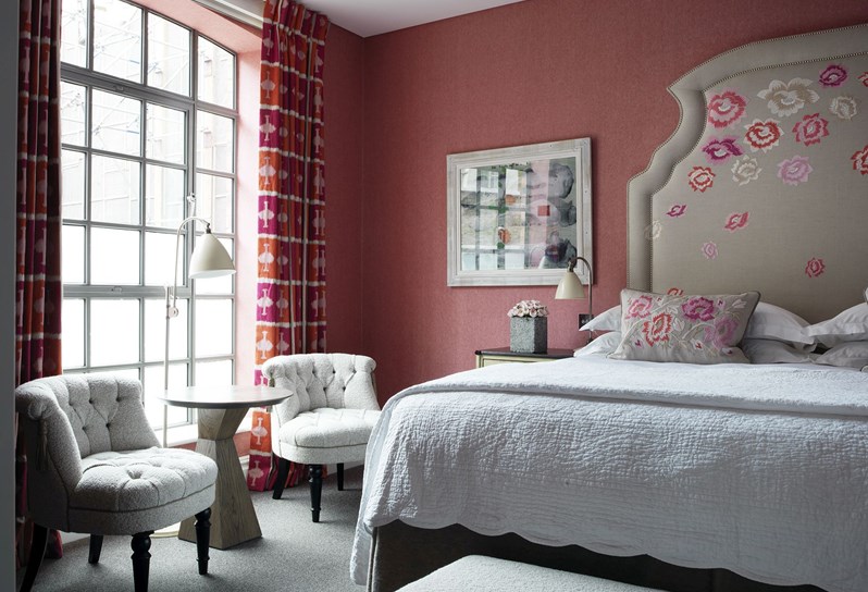 A closer look at a beautiful bedroom with a tan colored headboard with pink flower details on the headboard and the pillows, accented by the warm pink wall. Two textural chairs sit in front of the floor to ceiling windows and the curtains are patterned in pink, orange and white in Kit Kemp's own design.