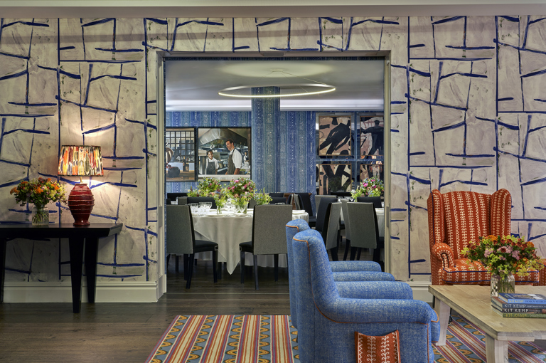 An image of all the Sandra Blow and Indigo private events rooms at The Soho Hotel. In the for ground of the image we see the back of two blue armchairs pointing towards the right, in the background we see through the open sliding doors into the Indigo Room which is set up with round tables with white tablecloths, placemats, plates, cutlery, glassware and flowers in the centre.  
