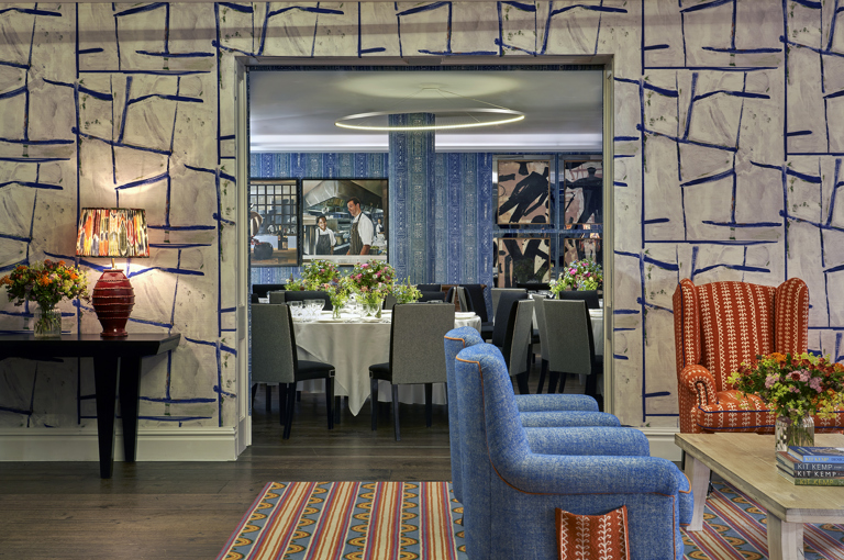 An image of all the Sandra Blow and Indigo private events rooms at The Soho Hotel. In the for ground of the image we see the back of two blue armchairs pointing towards the right, in the background we see through the open sliding doors into the Indigo Room which is set up with round tables with white tablecloths, placemats, plates, cutlery, glassware and flowers in the centre.  