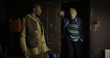 A still from the film "The Christophers": A women wearing a brown waistcoat looks over at an elderly man wearing a blue striped jumper
