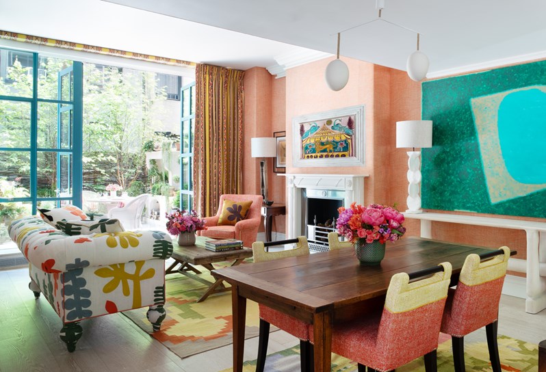 An open plan living/dining space with a wooden dining table and orange and yellow chairs. The walls are orange and there is a sofa upholstered in a multicoloured fabric. Bifold doors open onto a terrace with a table and chairs