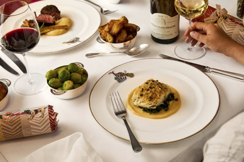 This image shows a festive Christmas meal in Brumus at Haymarket Hotel, with mythical creature crockery and dishes from the festive menu, including fish, steak, roast potatoes and Brussels sprouts, accompanied by red and white wine and Christmas crackers.