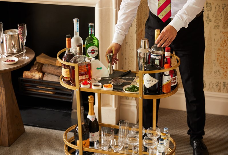 A gold framed cocktail trolley, a shelf at the bottom holds tumbler, highball and coup glasses and sparkling wine and tonic bottles. On the top tray there are different bottles of spirits, cocktail mixing kit, 3 mini bowls of dried fruit. A man is leaning down behind to make a cocktail.