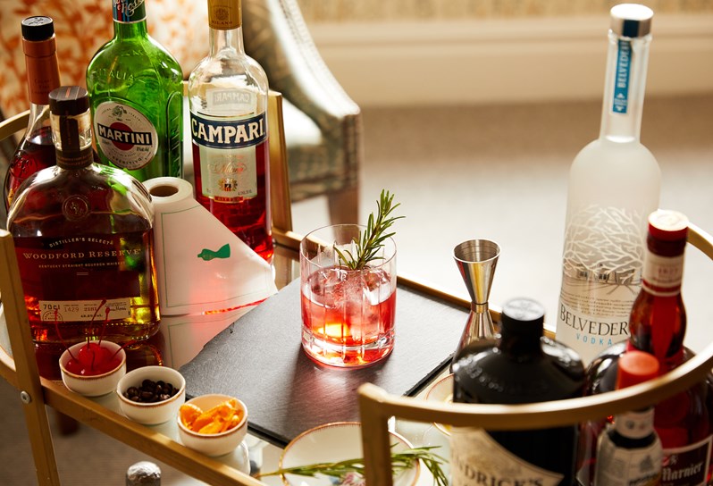 A gold framed cocktail trolley shot from above. On the top tray there are different bottles of spirits, cocktail mixing kit, 3 mini bowls of dried fruit and a negroni.