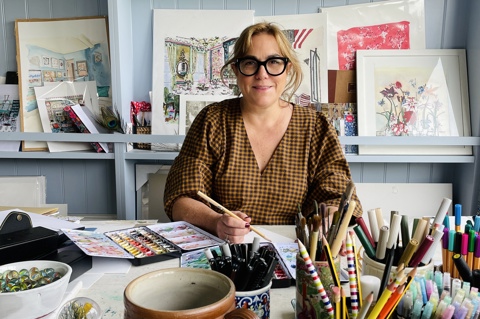 Artist SJ Axelby sits in her studio holding a paintbrush surrounded by pots of pens and pencils