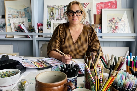 Artist SJ Axelby sits in her studio holding a paintbrush surrounded by pots of pens and pencils