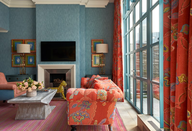 A living area. A blue and orange armchair in front of a coffee table opposite an orange 'Chainstitch' sofa which matches curtains on the windows behind. A fireplace, TV, a miniature wooden tiger statue and two chest of drawers either side with lamps on.
