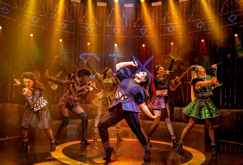 The six lead cast members of SIX the Musical. One woman stands in the centre, tilting her head back and holding a microphone above. Five other cast members surround her dancing and singing into microphones. All are dressed in shiny, structured dresses and wearing crowns.