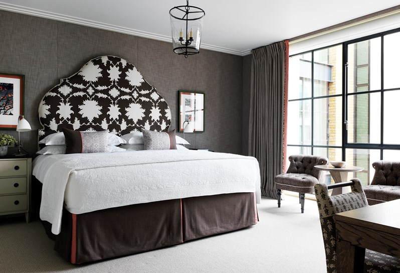 A light bedroom with king-size bed. Positioned in between two cream bedside tables with black tops, the bed has white linen and a large feature headboard, patterned in a bold black and white print. The bed rests against a neutral grey fabric wall with floor to ceiling windows to the right, framed by grey curtains with an earthy red trim.