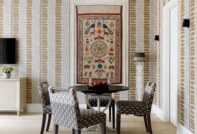 A view of the black circular dining table with four grey and cream patterned dining chairs. A colourful framed tapestry hangs on the wall behind the table with the walls decorated in a vertical stripe pattern in earthy tones.