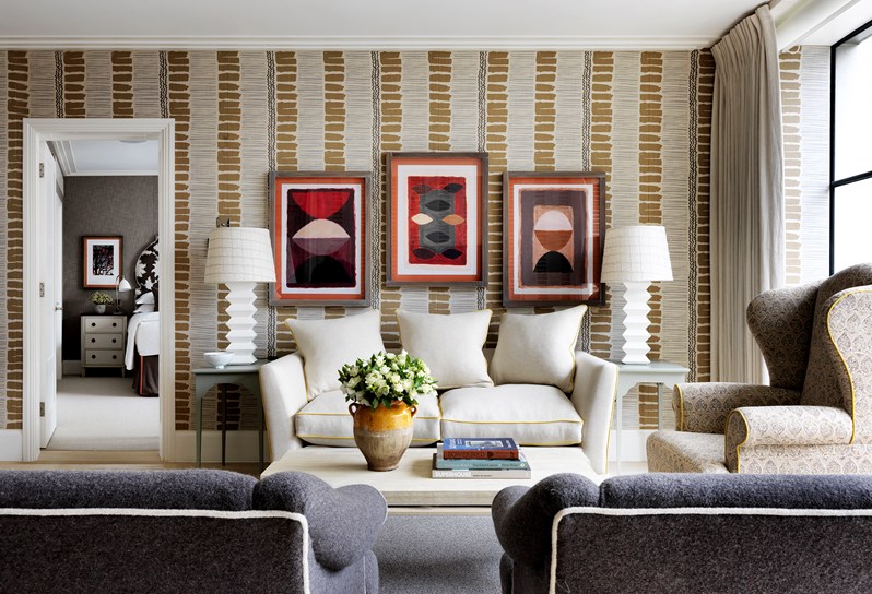A view of the living area. A cream sofa rests in front of a vertical striped wall with neutral and earthy tones. An oak coffee table sits in front of the sofa, with a cream and black wingback chair with a yellow trim directly to the right hand side in front of floor to ceiling windows. Two grey armchairs with white trim sit looking directly opposite the sofa and coffee table.