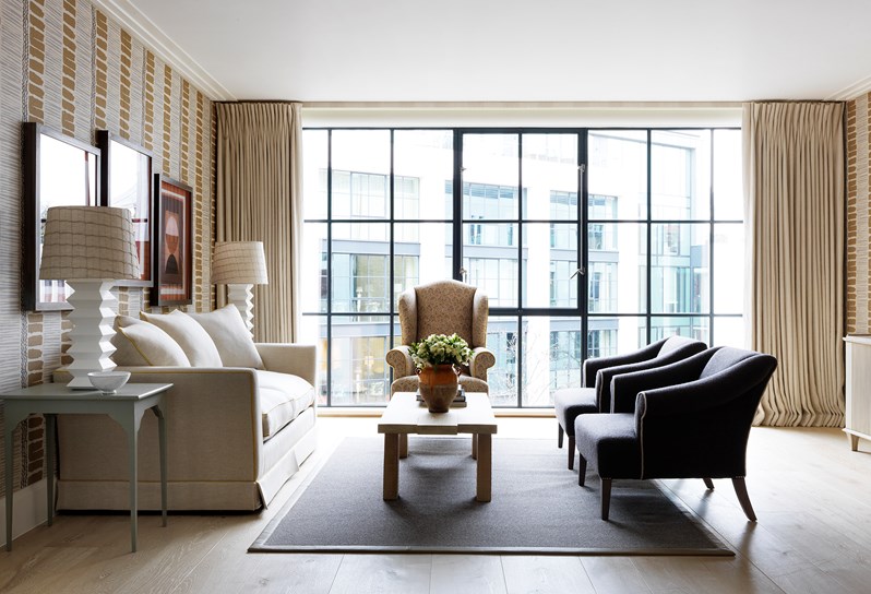 A side on view of the living area looking towards floor to ceiling windows, frame d by cream curtains with a cream and black wingback chair sitting directly in front. To the left, a cream sofa rests against a vertical stripe patterned wall, decorated in neutral and earthy tones. Opposite the sofa are two grey armchairs with an oak coffee table resting in between.