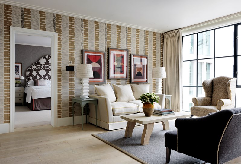 A angled view of the living space looking into the bedroom. A cream sofa rests in front of a vertical striped wall with neutral and earthy tones. An oak coffee table sits in front of the sofa, with a cream and black wingback chair with a yellow trim directly to the right hand side in front of floor to ceiling windows. An armchair with white trim sits looking directly opposite the sofa and coffee table.