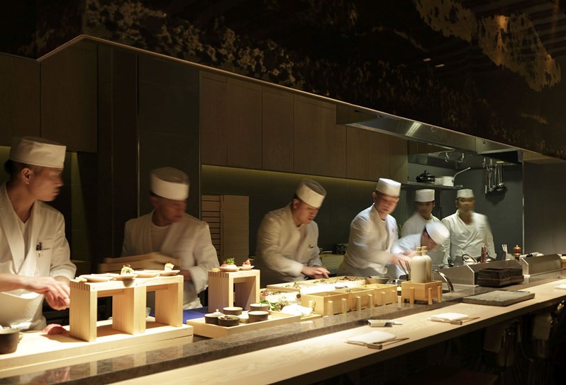 A side view of an open kitchen with seven chefs in white chef outfits, working at various stations.