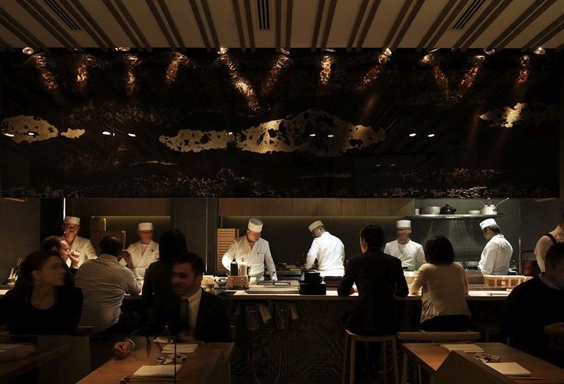 A busy restaurant with lots of diners and an open kitchen with chefs in white chef clothes working.