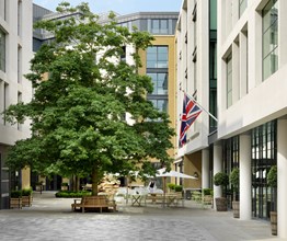 The entrance to Ham Yard Hotel with the British Flag leading the way in on the right, with the courtyard in front boasting leafy trees and alfresco dining.