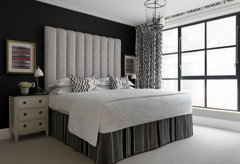 A light bedroom with king-size bed. Positioned in between two cream bedside tables with black tops, the bed has white linen and a large feature headboard, lightly patterned in cream and grey. There are floor to ceiling windows with black and white zig-zag patterned curtains.
