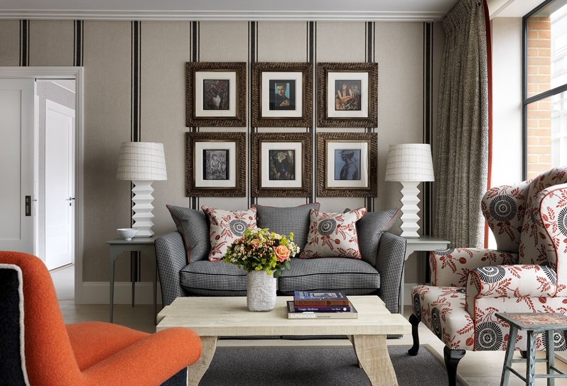 A view of the living area. A cream and black striped sofa rests in front of an earthy beige wall with a wide stripe pattern. An oak coffee table sits in front of the sofa, with a red, cream and black wingback chair directly to the right hand side in front of floor to ceiling windows, that are framed by black and white patterned curtains. In the front left of the picture, an orange armchair sits directly opposite the sofa and coffee table.