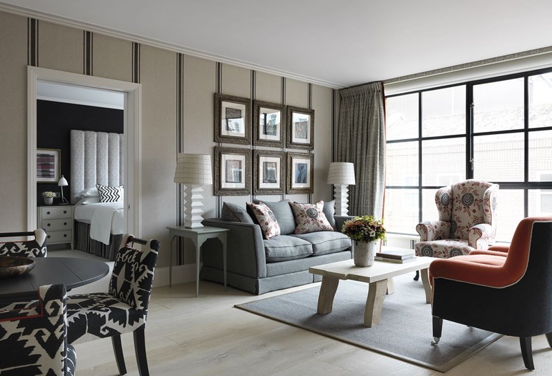 A wide angle view of the living space, with a view into the bedroom. A cream and black striped sofa rests in front of an earthy beige wall with a wide stripe pattern. An orange upholstered armchair sits opposite the sofa with an oak coffee table resting in between. To the right are floor to ceiling windows, framed by black and white patterned curtains with a cream, red and black wingback chair in front. To the left of frame is a circular black dining table with four black and white patterned chairs.