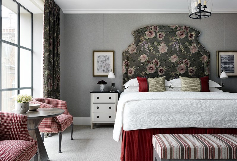 A light and bright bedroom with king-size bed. The bed has white linen and a large feature headboard with a floral pattern in olive green and soft pinks. At the foot of the bed is a cream, red and black striped footstool, and sitting to the left of the bed is a cream bedside table with black top. There are floor to ceiling windows to the left with curtains matching the patterned headboard. In front of the window is an occasional table, with two red and white patterned armchairs on either side.