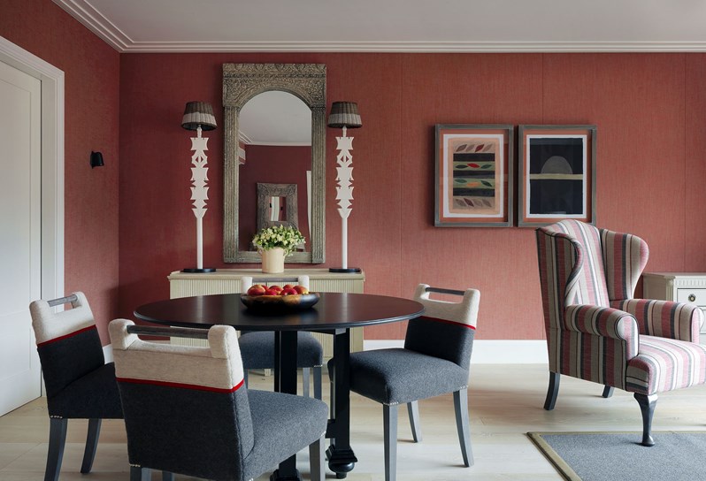 A black circular dining table sits encircled by four grey and cream dining chairs. To the right sits a red, white and cream striped wingback chair, resting on a black and white rectangular rug with an olive green trim. The walls are furnished with earthy red fabric with a sideboard resting by the wall towards the back left corner. Upon the sideboard are two tall lamps, either side of a long wooden framed mirror.