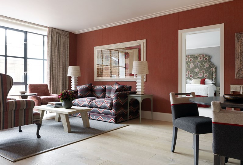 A wide angle view of the living space, with a view into the bedroom. A grey, red and cream zig zag patterned sofa rests in front of an earthy red fabric wall. A red, white and cream striped wingback chair sits opposite the sofa with a coffee table resting in between. To the left are floor to ceiling windows, framed by cream patterned curtains with a cream and red armchair in front. To the right of frame is a circular black dining table with four grey and cream patterned chairs.