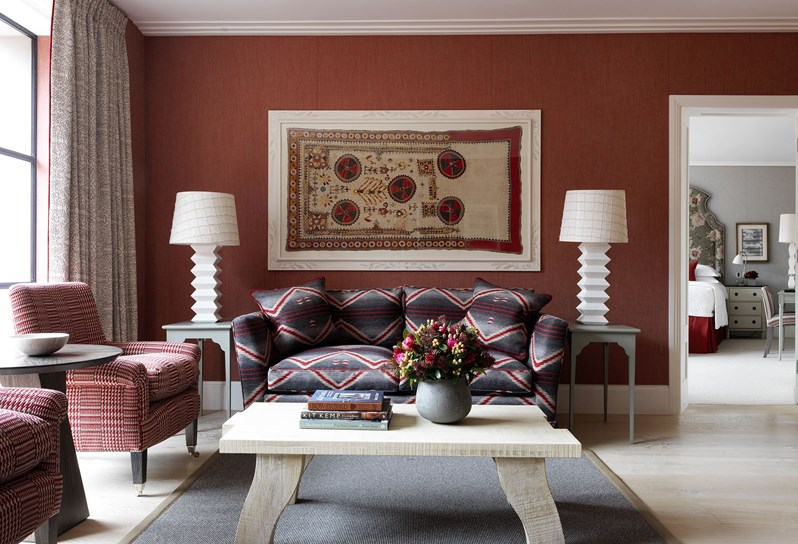 A straight on view of the living space looking into the bedroom. A grey, red and cream zig zag patterned sofa rests in front of an earthy red fabric wall. To the left are floor to ceiling windows, framed by cream patterned curtains with a pair of cream and red armchairs in front and an occasional table sat in between. In the centre is an oak coffee table sitting atop a black and white patterned rectangular rug, with an olive green trim.
