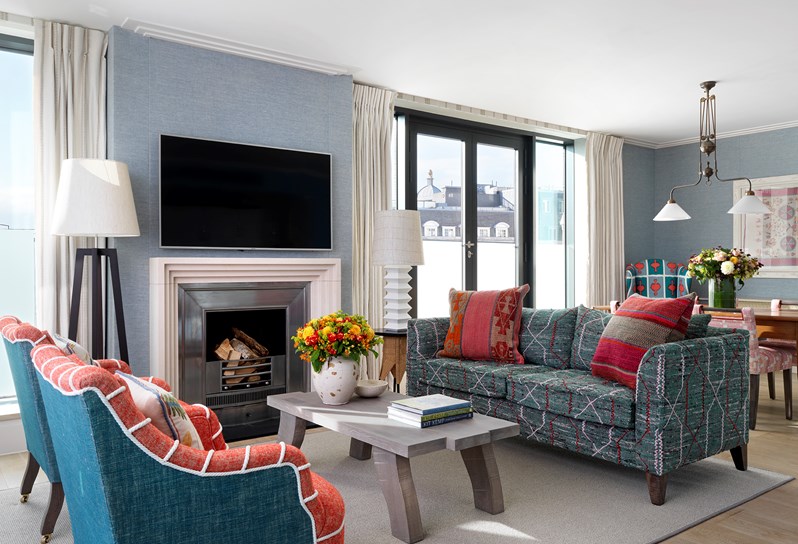 An angled view of the light and bright living area on the upper floor of the duplex apartment, with floor to ceiling windows lining two perpendicular walls. At the front is a circular dining room table with four green patterned dining chairs with a wingback floral chair resting on the right, in front of the windows. A long occasional table rests against the blue sofa, opposite two red patterned armchairs.