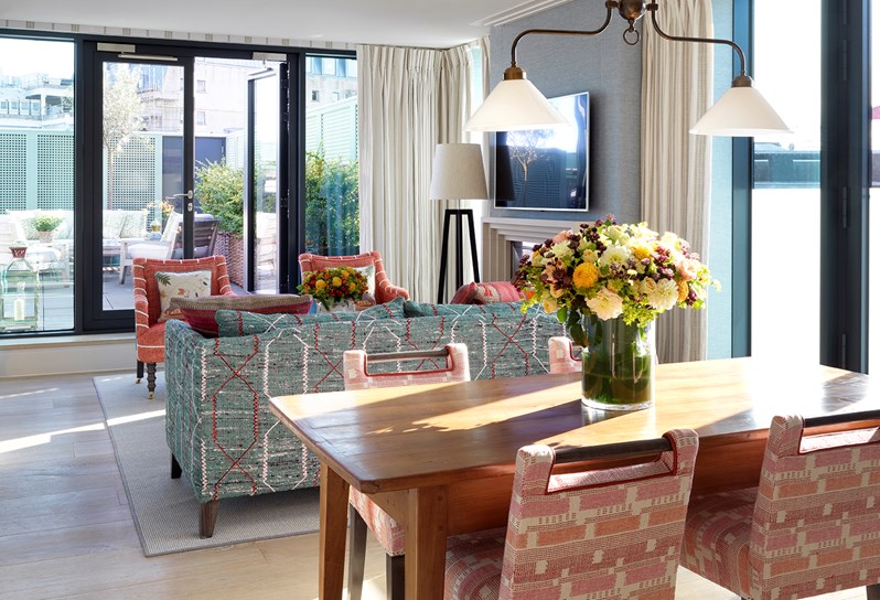 An angled view of the light and bright living area on the upper floor of the duplex apartment, with floor to ceiling windows. At the front is a dining room table with four red patterned dining chairs. To the left is a low dividing wall, marking one side of the staircase leading up from the lower floor. A blue sofa sits opposite two red patterned armchairs, resting in front of the windows.