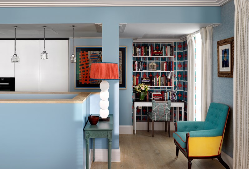 A view over the dividing walls that enclose the staircase leading up from the lower floor of the duplex apartment.  Looking towards the back wall of the kitchen, are long white cupboards with an inbuilt oven and to the right, an occasional table with a tall white lamp on top.