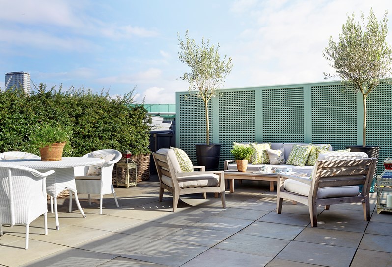 A view of the large terrace with skyline views across London. The edge of the terrace is lined with wicker wooden fencing in front of a green hedge. On the right hand side of the terrace is a white wicker circular dining table with four white chairs and to the left is a white wicker deck chair.