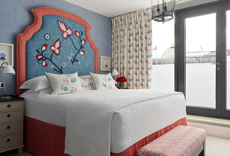 One of three bedrooms in the duplex apartment. A king-size bed is positioned in between two cream bedside tables with black tops, the bed has white linen and a blue floral patterned feature headboard. The bed has a red fabric footstool at the end and the headboard rests against an blue fabric wall, with floor to ceiling windows and a balcony door to the right.