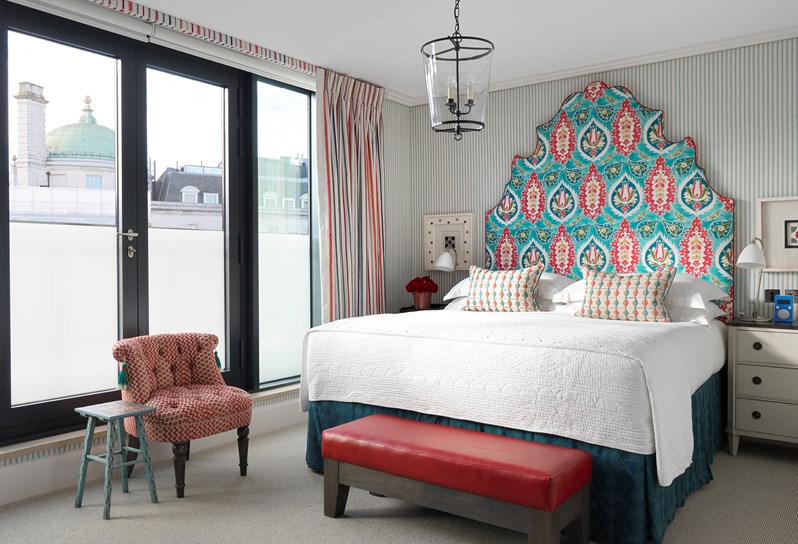 A king-size bed with white linen and a turquoise patterned feature headboard. A cream bed side table with a black top sits to the right hand side of the bed, resting up against an striped fabric wall. To the right are striped curtains.