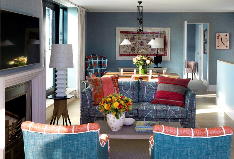 A view of the living area leading into the bedroom.  An oak coffee table sits in between a blue sofa and two red patterned armchairs. A low wall on the right hand side, divides the kitchen area from the living space with a staircase leading up from the lower floor. Behind the sofa is a dining table, sitting next to the left hand wall lined with floor to ceiling windows and doors leading to a balcony.
