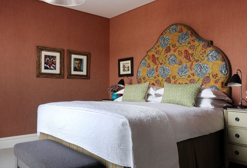 View of a bed with a headboard in rich shades of orange, red and blue. The walls are a coordinating deep red and there are three pieces of framed art on the far wall.