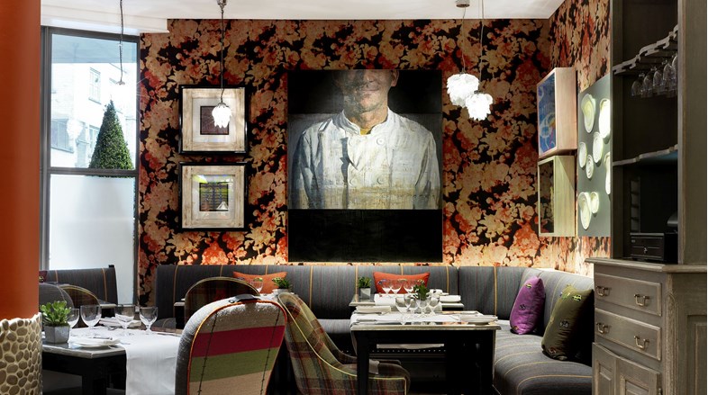 A photograph of refuel restaurant. Table covered in white table cloths surrounded by grey bench seating. The walls are clad in an orange floral wall paper. There is a painting of a chef on the wall.