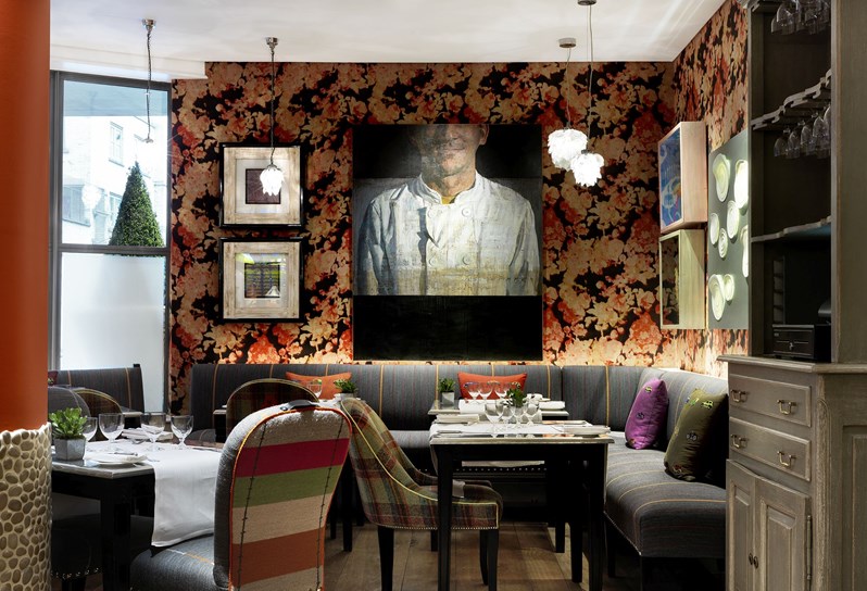 A photograph of refuel restaurant. Table covered in white table cloths surrounded by grey bench seating. The walls are clad in an orange floral wall paper. There is a painting of a chef on the wall.