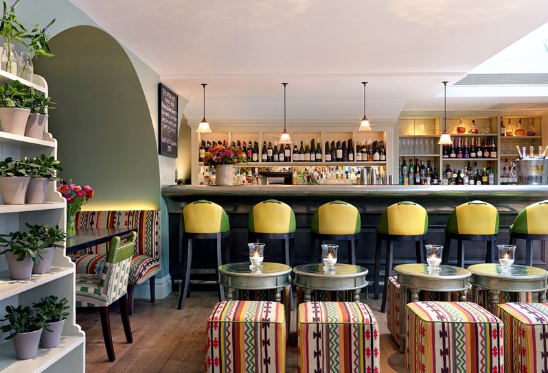 Colourful details in low lounge tables, seating puffs and comfortable chairs in the seating area of The Potting Shed Bar, facing the bar with five bold yellow and green high chairs.