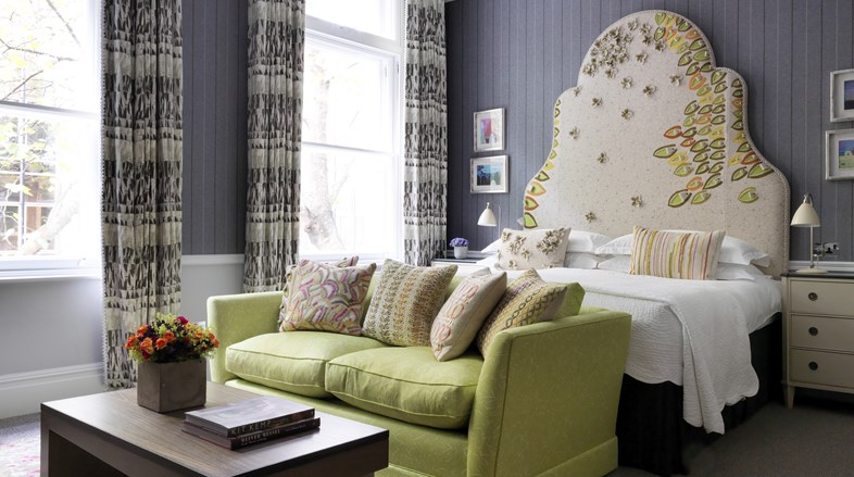 Image of a spacious and bright bedroom, with grey wallpaper and beautiful pattern curtains and headboard, the room has a king sized bed and a wooden coffee table with a colourful flower arrangement