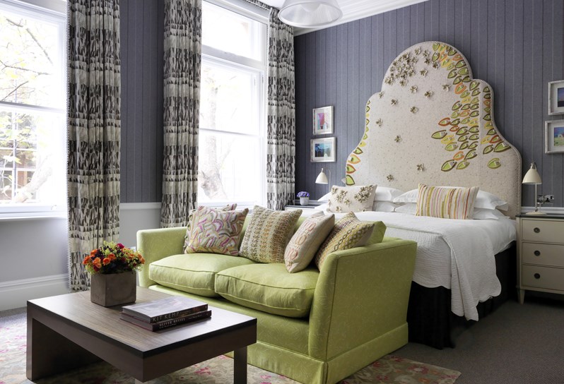 Image of a spacious and bright bedroom, with grey wallpaper and beautiful pattern curtains and headboard, the room has a king sized bed and a wooden coffee table with a colourful flower arrangement