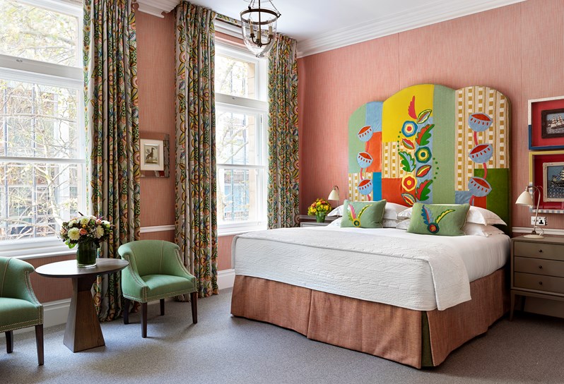 Room has a pink wallpaper with a bold colourful headboard with two big windows.