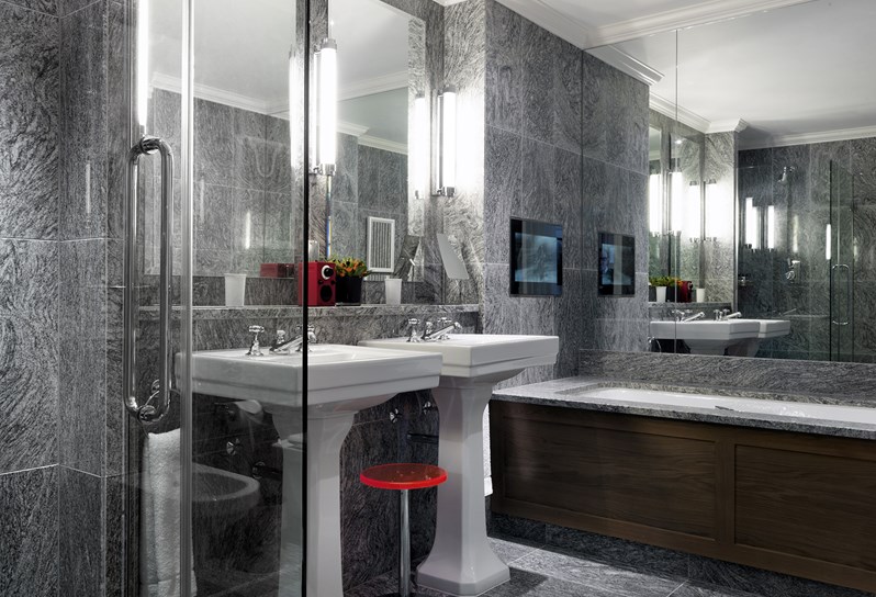 A beautiful granite bathroom with walk in shower to the left, two wash basins in the center and bathtub and television in the front.