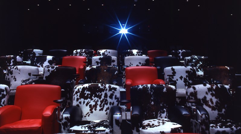 The screening room at Soho Hotel.  Cinema style red leather chairs are interspersed with black and white cowhide chairs. In the background is a projector pointing towards the screen.