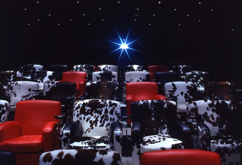 The screening room at Soho Hotel.  Cinema style red leather chairs are interspersed with black and white cowhide chairs. In the background is a projector pointing towards the screen.
