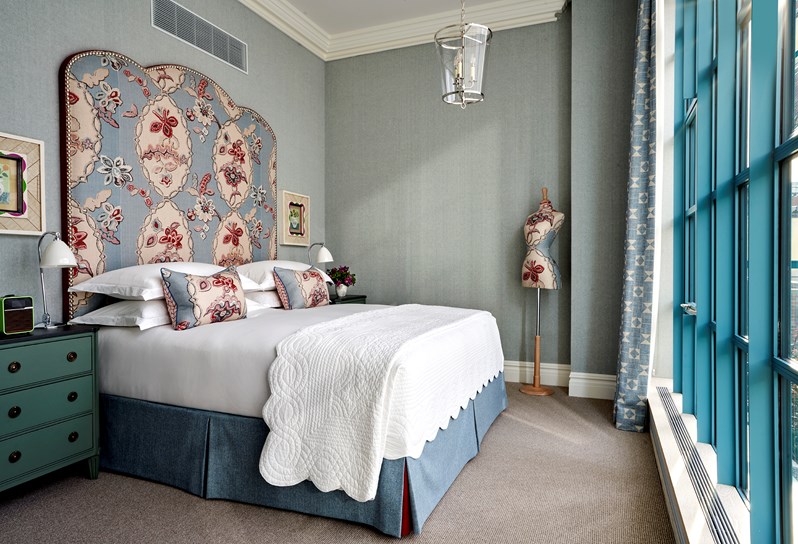 A blue colour scheme bedroom. In the centre, facing the floor to ceiling windows, there is a bed with a statement headboard in blue 'Chainstitch' fabric which matches the cushions and the mannequin in the right hand corner. There are turquoise bedside tables on either side of the bed.